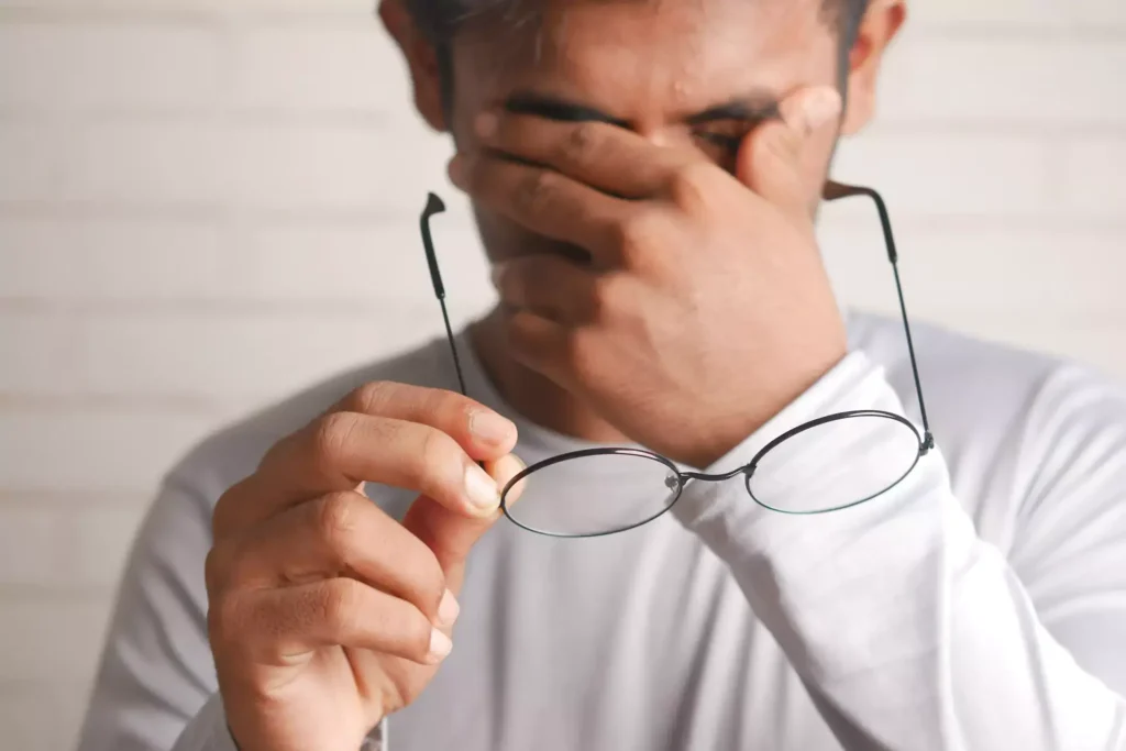 Person with white shirt holding glasses in one hand and rubbing eyes with the other hand